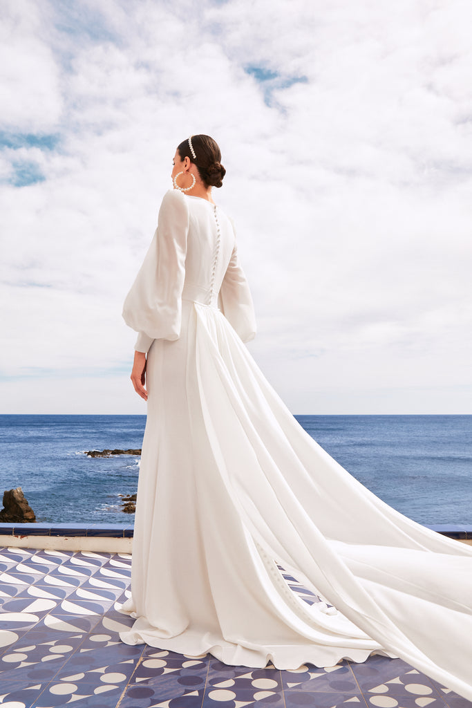 The model is facing away from the camera to show the buttons going down the back of the dress to the drop waist. Also shown is the small train and overtrain leaving from where the buttons stop. The puffy sleeves show some of the models arm though its translucent material.