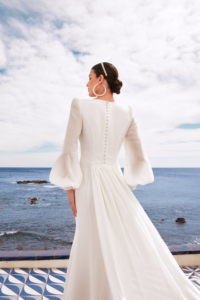 The torso of the back of the model highlighting the button details going down the back, the bishop sleeves, and the pearl hoop earrings and headband. The sky and ocean are shown in the background.